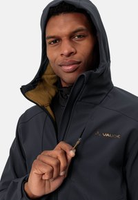 Męska ciemnoszara kurtka softshell z zapięciem na zamek przy kołnierzu, brązowym polarem wewnątrz i złotym logo. Widoczna miękka tekstura oraz ergonomiczny krój.