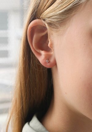 Zilveren stud oorbel met een klein rond gezicht, met een heldere edelsteen in het midden. Close-up afbeelding met een lichte huidtoon en lang haar.