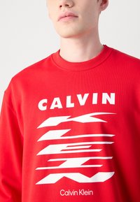 Sarkans džemperis ar baltu uzrakstu "CALVIN" un abstraktu grafisko dizainu. Ribota apaļā kakla izgriezums un mīksta, gluda auduma tekstūra.