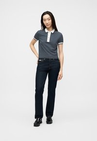 Γυναίκα που στέκει με το ένα χέρι στη μέση, φορώντας μπλε μπλουζάκι πόλο με ρίγες, σκούρο τζιν και μαύρα γυαλιστερά παπούτσια σε απλό φόντο.