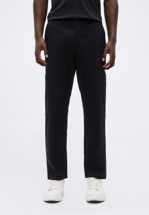 Mann iført svarte bukser med rette ben og hvite joggesko, står med hendene avslappet langs siden mot hvit bakgrunn.