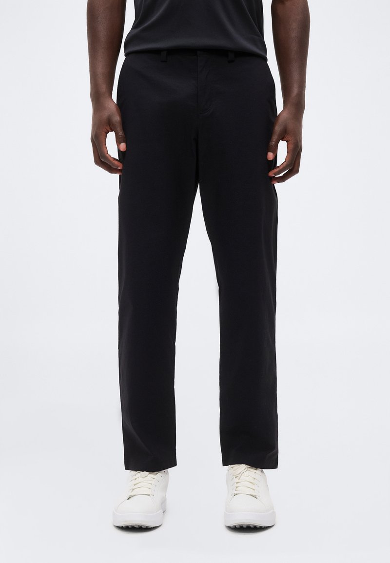 Homme portant un pantalon noir à jambes droites et des baskets blanches, debout les mains détendues le long du corps sur fond blanc.