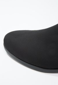 Punta de zapato de ante negro con forma suave y redondeada y suela delgada de goma negra sobre un fondo blanco.