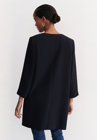 Dunkelblauer, knielanger Mantel mit geradem Schnitt, Drei-Viertel-Ärmeln und minimalistischem Design. Strukturierten Stoff und klare Linien.