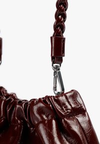 Detalle de bolsa de cuero burdeos con una correa trenzada, herrajes de plata y textura recogida en el cuerpo principal.