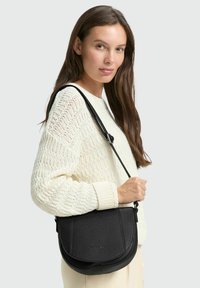 Borsa a tracolla in pelle nera con forma arrotondata, superficie testurizzata e tracolla regolabile, tenuta da una persona con un maglione crema.