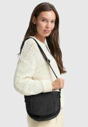 Borsa a tracolla in pelle nera con forma arrotondata, superficie testurizzata e tracolla regolabile, tenuta da una persona con un maglione crema.