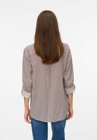 Femme aux longs cheveux bruns de dos, portant une chemise blanche à manches longues avec des rayures verticales brunes et un jean bleu.