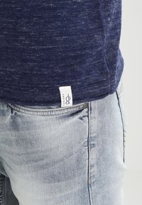 T-shirt bleu marine texturé avec une petite étiquette blanche à l'ourlet, associée à un jean en denim bleu clair présentant des zones décolorées et des coutures visibles.