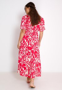 Maxi jurk in rood en wit bloemmotief, korte mouwen, geplooide taille, vloeiende stof, gedragen met lichtgekleurde sandalen.