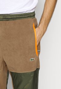 Bruna fleecebyxor med en elastisk grön midja, orange och blå dragkedjefickor samt en liten grön logotyp. Strukturerad, avslappnad design.