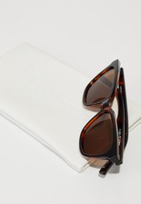 Skilpadde-sunglasses med brune linser, med vinklede rammer. Følger med en hvit, teksturert beskyttelsesetui preget med "Le Specs."