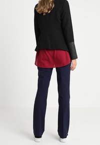 Svart skräddarsydd kavaj med läderdetaljer, matchad med en vinröd blus, marinblå byxor och vita sneakers. Bakifrån visas rena linjer och passform.