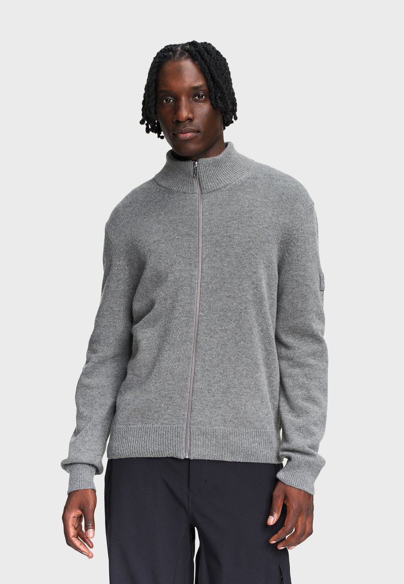 Man met kort gedraaid haar, gekleed in een grijze gebreide vest met rits en zwarte broek, staand tegen een effen lichte achtergrond.