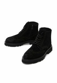 Botines de ante negro con detalles de patrones brogue, que presentan una suela gruesa negra y un diseño de cordones, acompañados de un cierre con cremallera en el lateral.