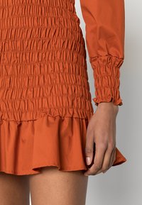 Vestido cor laranja enferrujada com textura franzida, mangas longas e bainha com volantes. O tecido parece suave, leve e ligeiramente brilhante.