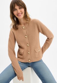 Lächelnde Frau in beigem Strickcardigan mit goldenen Knöpfen und blauen Jeans, sitzend mit ruhenden Händen, vor weißem Hintergrund.