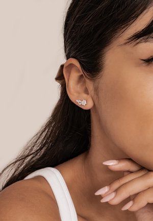 Woman’s left ear with silver floral-shaped stud earring and hand resting on chin, showing manicured nails and smooth skin.