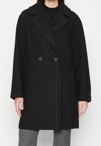 Cappotto in lana nera con design doppiopetto, ampi revers, dettagli di cucitura bianca a contrasto e tasche laterali. Presenta due bottoni neri.