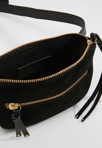 Musta mokkanahkainen olkalaukku kultaisilla metalliosilla, jossa on vetoketjullinen pääosasto ja etutasku, korostettuna nahkaisilla vetoketjenvetimillä.