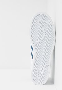 White athletic shoe with a textured rubber sole, featuring wavy patterns and dark blue stripes on the sides.