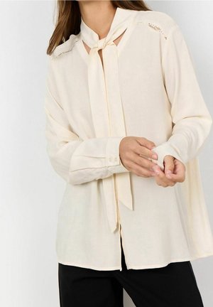 Blusa color crema con scollo annodato, maniche lunghe, polsini con bottoni e dettagli in pizzo sulle spalle. Tessuto leggero e dalla texture liscia.