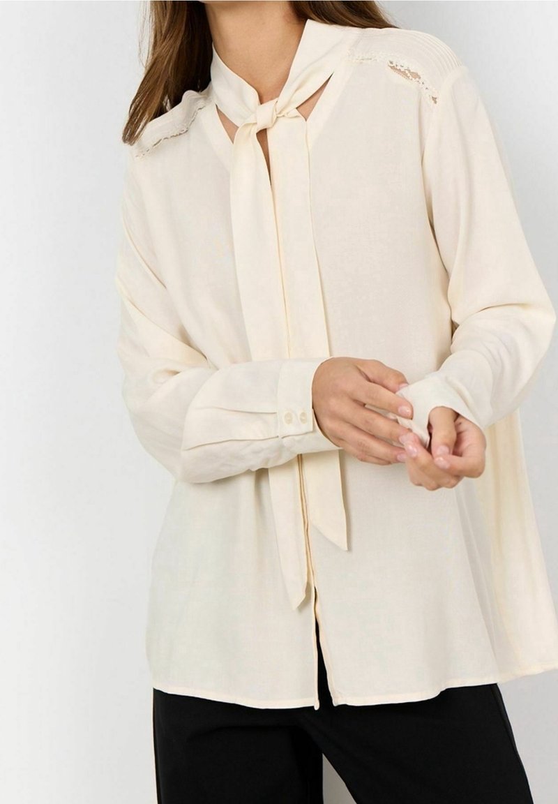 Blouse crème avec un col noué, manches longues, poignets à boutons et détails en dentelle aux épaules. Texture légère et lisse.