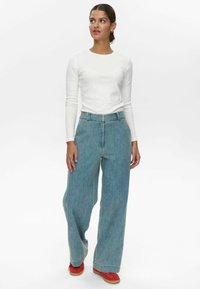 Könnyű, hosszúujjú fehér felső egy magas derekú, bő szárú kék farmernadrággal és piros tornacipővel párosítva, letisztult dizájnnal és minimális részletezéssel.