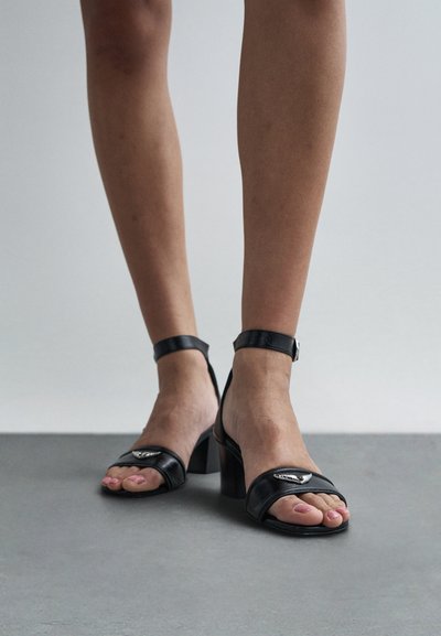 Čierne kožené sandále na vysokom podpätku s remienkom okolo členka, otvorenou špičkou a kovovým doplnkom na remienku. Textúrovaný povrch a široký podpätok.