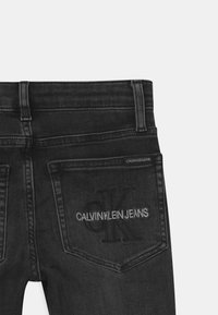 Jeans de mezclilla negra con un bolsillo trasero que presenta un logo en relieve "CK" y el texto "CALVIN KLEIN JEANS". Detalles de costura sutiles en todo el diseño.