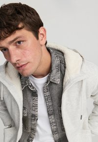 Giacca zip grigia chiara con cappuccio in felpa bianca testurizzata, sovrapposta a una camicia di jeans nera, indossata sopra una maglietta bianca.