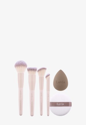 Cinq pinceaux de maquillage beiges avec des poils violet clair, une éponge marron en forme de goutte, et une houppette ronde avec le logo "Luvia".