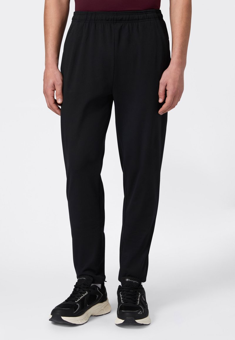 Man in zwarte elastische taillebandbroek en zwarte sportschoenen staat voor een witte achtergrond.