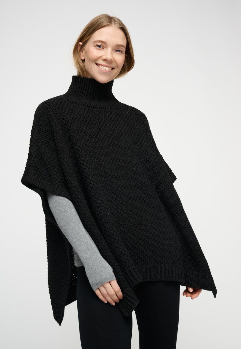 Svart stickad poncho med hög polokrage, med ett strukturerat mönster och ribbade kanter, kombinerad med en grå långärmad tröja under.