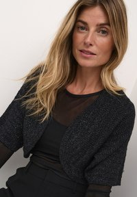 Black cropped jacket with short sleeves, textured fabric with metallic thread, worn over a sheer black top. Simple, rounded collar design.