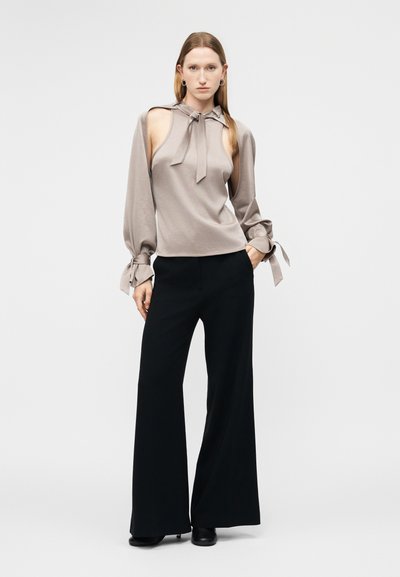 Femme portant un haut sans manches taupe avec un col noué en forme de nœud et de longues manches détachées, associé à un pantalon noir à jambes larges et des chaussures noires.