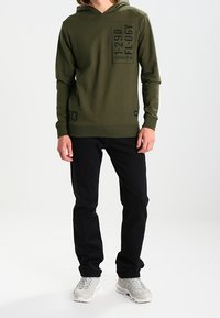 Oliivinvihreä huppari, jossa tasku ja tumma painettu teksti, yhdistettynä mustiin housuihin ja vaaleanharmaisiin lenkkareihin. Yksinkertainen muotoilu ja rento istuvuus.