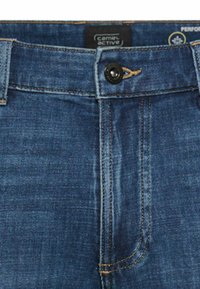 Voorste taillegebied van een blauwe spijkerbroek met zwarte knoop, beige stiksels en Camel Active merklabel aan de binnenkant van de tailleband.