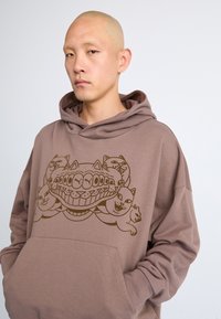 Brun hættebluse lavet af blødt stof, med et grafisk design af tegneserie-katte med overdrevede ansigtstræk. Inkluderer en brystlomme og afslappet pasform.