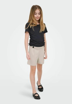 Young girl with long hair wearing a black ruched t-shirt, beige shorts with buttons, and black Mary Jane shoes, standing against white background.