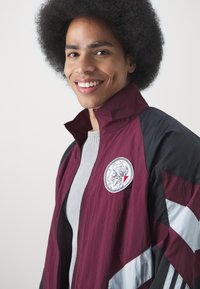 Corta-vento marrom e preto com um emblema redondo no lado esquerdo do peito, apresentado com riscas brancas nas mangas, tecido leve e gola com fecho éclair.