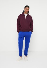 Mørkerød hættetrøje med lynlås og høj krave, parret med blå sweatpants og lyse slip-on sko. Glat stof, afslappet pasform.