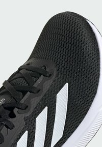 Schwarzer Sportschuh mit einem strukturierten Mesh-Obermaterial, weißen drei Streifen Akzenten und einer weißen Gummisohle. Verfügt über schwarze Schnürsenkel und ein schlankes Design.