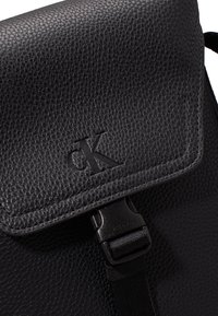 Mochila de cuero negro texturizado con cierre de solapa magnético, que presenta un logotipo en relieve y una correa ajustable.