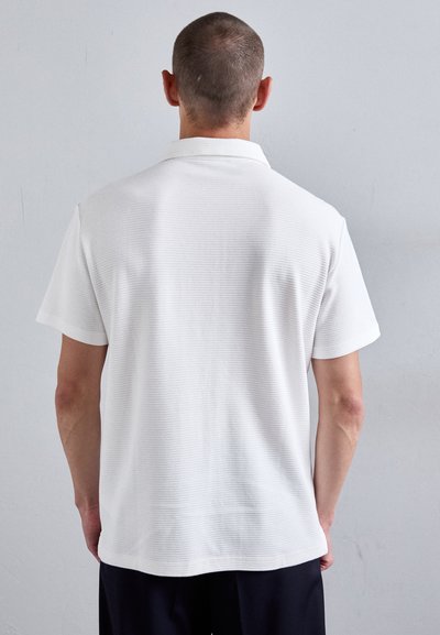 Homme avec les cheveux courts portant un polo blanc à manches courtes et un pantalon noir, se tenant face à un mur gris clair uni.