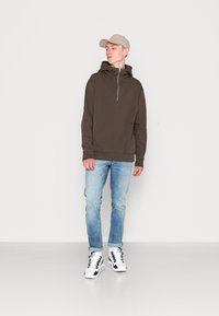 Brauner Zip-Hoodie mit hohem Kragen, hellblaue Jeans und schwarz-weiße Sneakers, ergänzt durch eine hellbeige Baseballkappe.