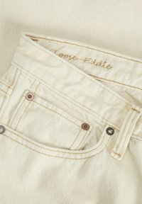 Cremefarbene Jeans in lockerer Passform mit sichtbaren Nähten, einer kleinen Vorderntasche und metallischen Knöpfen am Bund. Dezente Texturdetails.