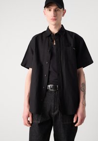 Camisa negra de manga corta con botones y bolsillo, combinada con una camiseta sin mangas negra y pantalones vaqueros oscuros. Presenta un diseño simple y una textura suave.