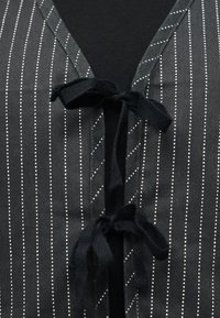 Μαύρο ένδυμα με λευκές κάθετες ριγέ τελείες που διαθέτει δύο μαύρες υφασμάτινες κορδέλες δεμένες σε κόμπο στο κέντρο του μπροστινού μέρους στο λαιμό.