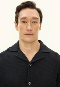 Czarna teksturowana koszula z kołnierzem typu camp, zapinana na guziki z przodu, z dwoma widocznymi guzikami. Tkanina ma subtelny wzór.